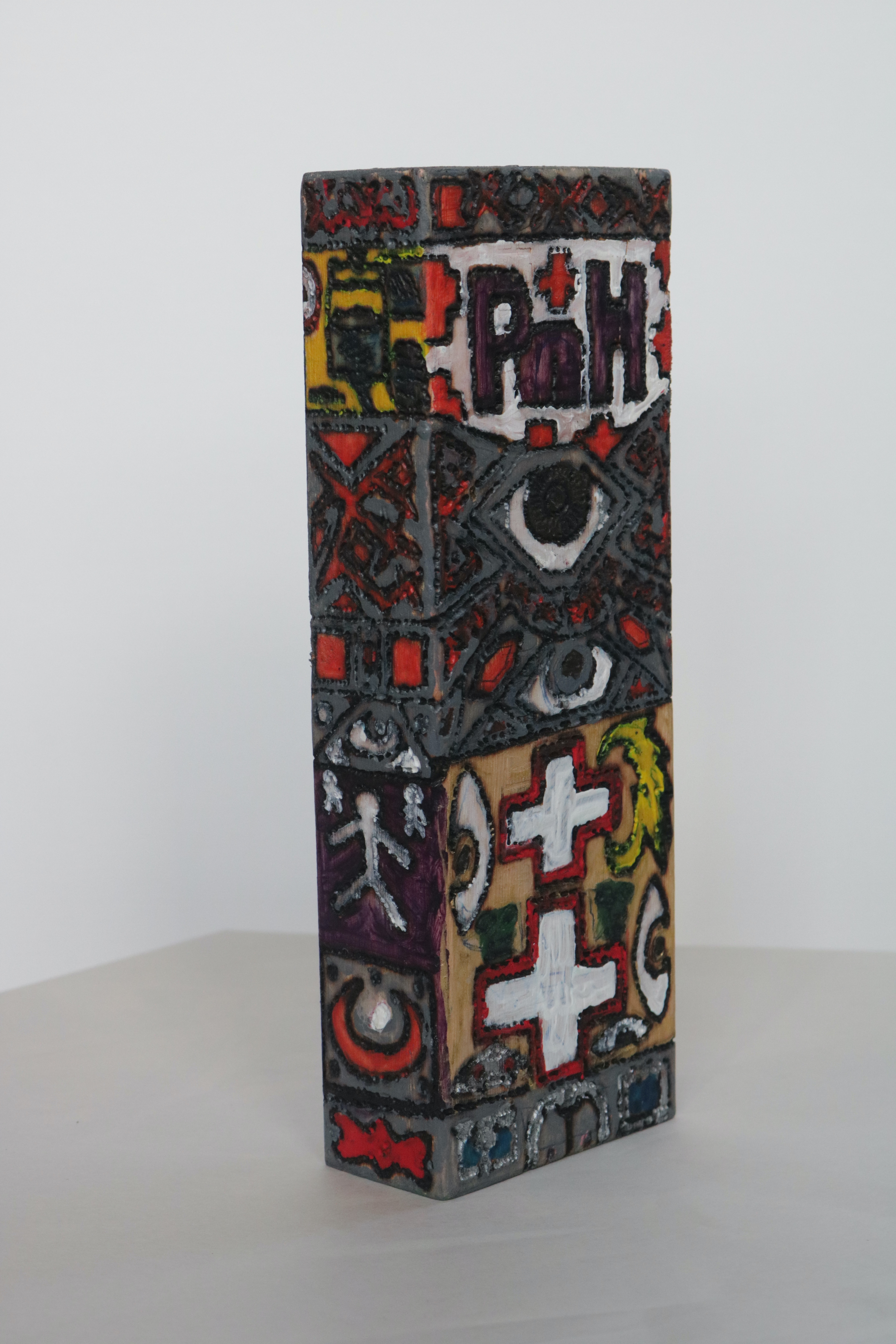 A slim, tall, wooden block. The letters P n H burnt in and painted. There are crescents, crosses, and eyes, burnt throughout the cluttered patterns.