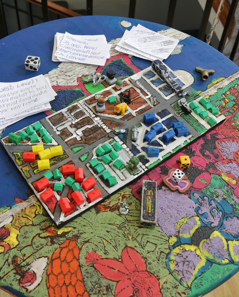 the board is on a table decorated by Jonny, but obscures the details. The board is painted with streets and parks. It is covered in plastic monopoly houses. There are action cards on the table; one says asteroids.