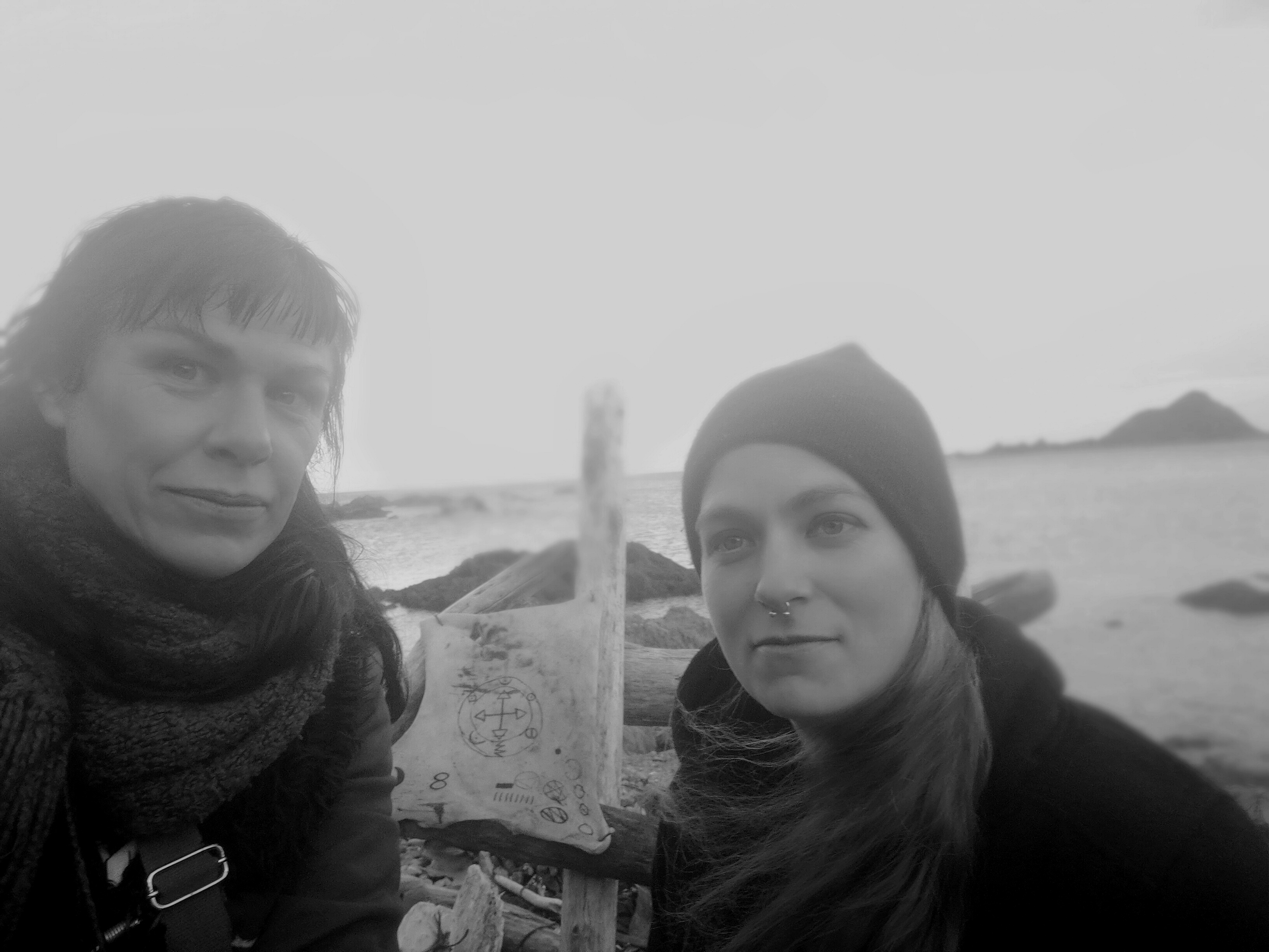 Holly and Greta stand at island bay. There is a pile of driftwood that has a piece of leather nailed to it. the leather looks like rawhide and is covered in magical sygils.