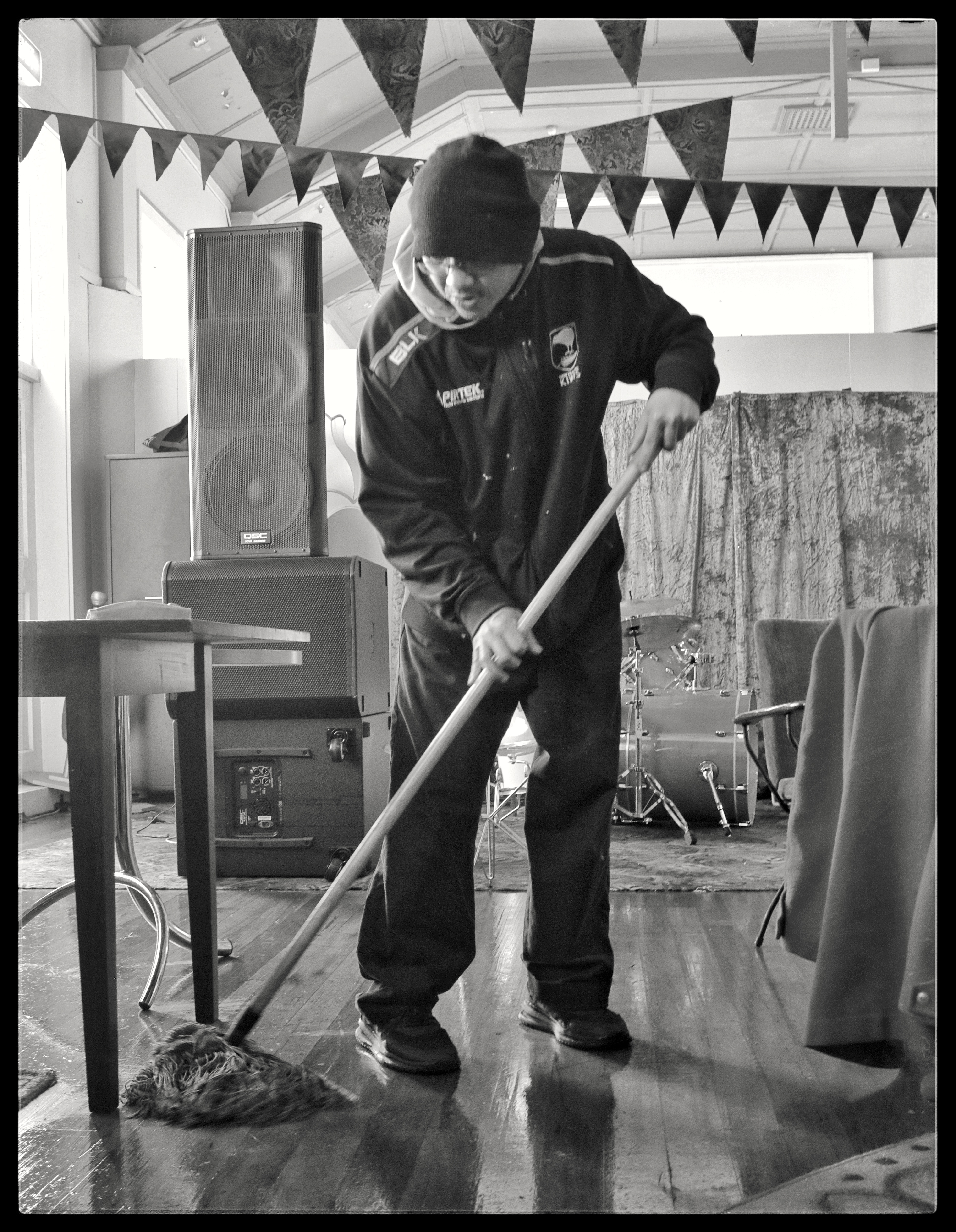 A black and white photo of Jonny Kan mopping the floor, the large speaker of the Snails PA is in the background
