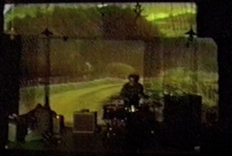 A lo-res photo of a person at the drumkit, there is a grainy projection as lighting on a haphazard screen.