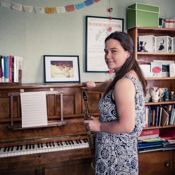 Jasmine standing in front of an an old upright piano. It is a very doemnstic setting with photos on top of the piano and a pleasingly unitdy bookshelf in the background.