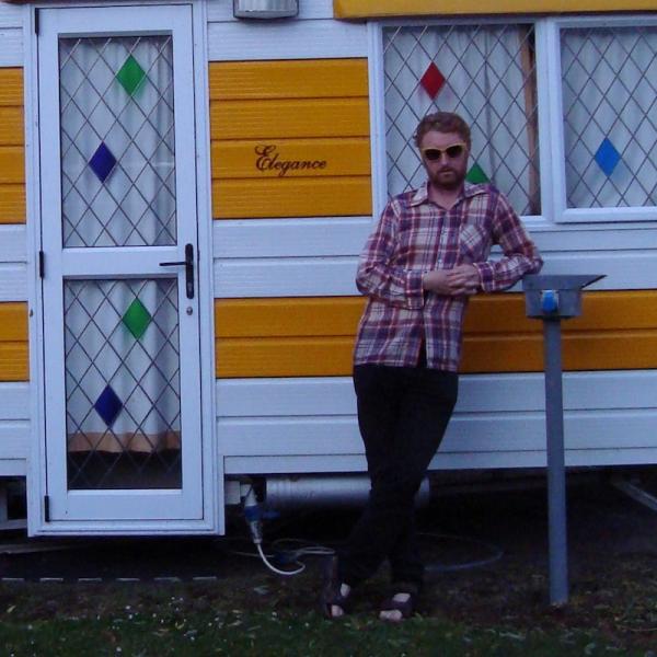 Mark is leaning nonchanontly in front of a happy yellow and white caravan