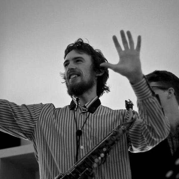 A back and white photo of Jake holding his hands up in a gesture of conducting some musicians unseen. There is a music stand to one side of him.