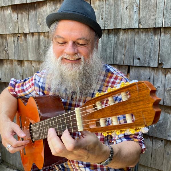 Chris has a large grey beard and is wearing a small pork pie hat. He is playing a nylon string guitar and is smiling with a large joyful smile.