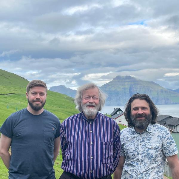 Three bearded men stand in the green jagged hills of the Faroe Islands