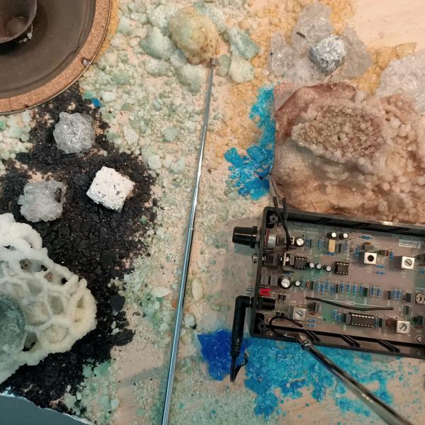 A table with whote a blue (copper?) crystals growing on it. There is a raw speaker cone and an open electronic device.