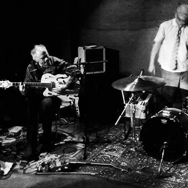 Black and white photo of Greg Malcolm hunched over his guitar and pedals and a blurry Stefan Neville looming over his drumset