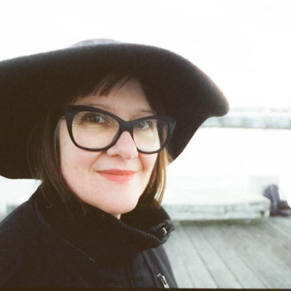 Sonya Waters smiles at the camera in a black hat and black outfit on a pier on the sea