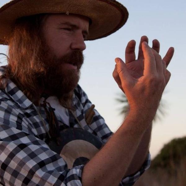 Chris O'Connor stands in front of a sunset explaining something with his hands. He's wearing a large brim hat