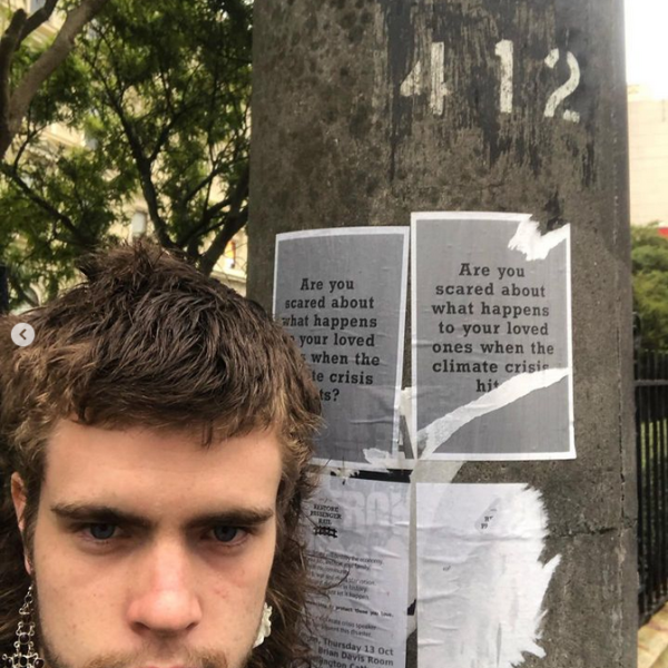 A photo of Baxter in front of a lamp post. The pole has flyers stuck to it, they are slightly torn, and they read "are you ascred about what happens to your loved ones with the climate crisis hits?". Baxter has a mullet and elaborate dangly earrings