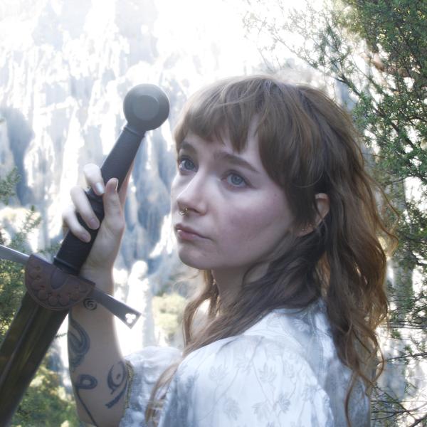 A glowy photograph of Fox Moss posing with a sword in front of cliffs looking in the distance. 