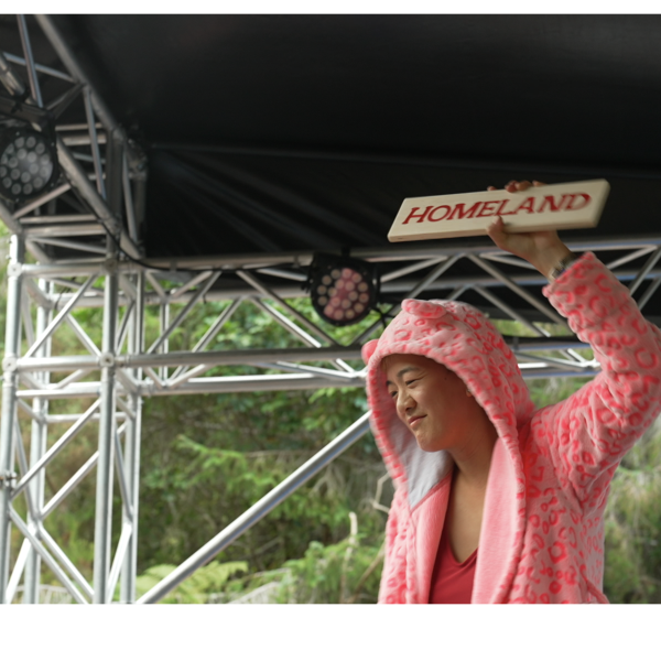 Kristen is on an outdoor stage at Camp a Low Hum. The bush is behind her as she holds up a sign that reads Homeland