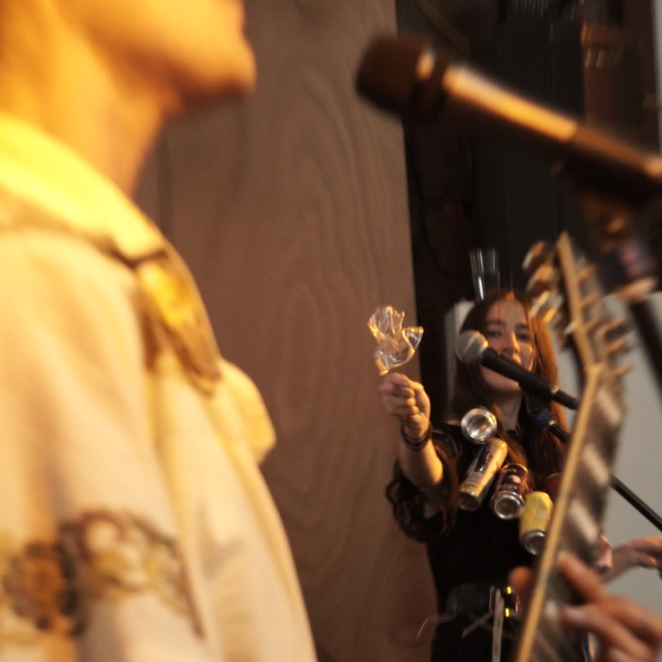 In the foreground is someone playing an electric guitar. In the background someone is playing junk percussion with joy and gusto. The percussion looks to be plastic cups and aluminium cans.