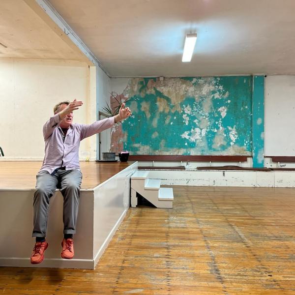 Stephen sits on the corner of a stage at a community hall. The wooden floors are well kept but are starting to show the years of use. The walls are in a state of unkempt life.