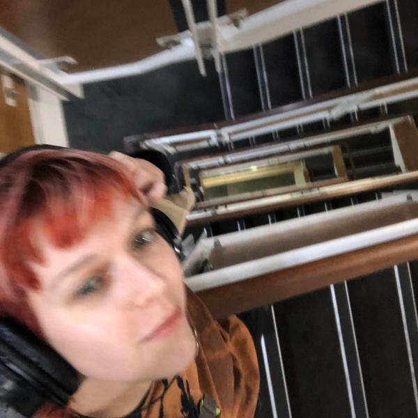 Tash has headphones on and is looking up toward the camera above them. They are at the top of a large stairwell