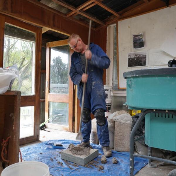 Tim smashing lime stobe with a pole, preparing to make a Roman wall. he is weraing blue overalls, saftey goggles, and is covered in dust.