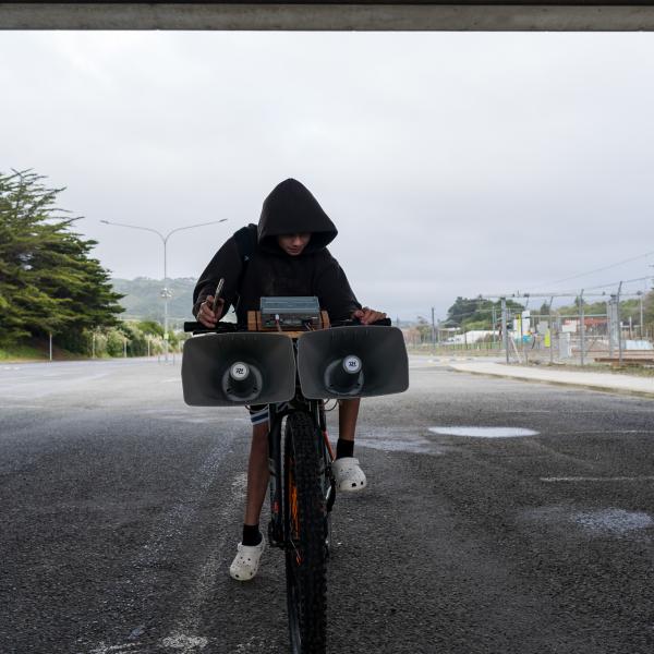 A kid on their bike by the train tracks. The kid is holding a phone and tweaking some knobs on some audio gear mounted on the handle bars. There are two huge siren speakers hanging off the front of the bars.
