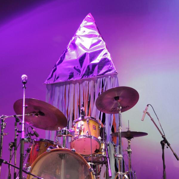 Andy playing the drums in a large silver pyramid that hides his entire body and head.