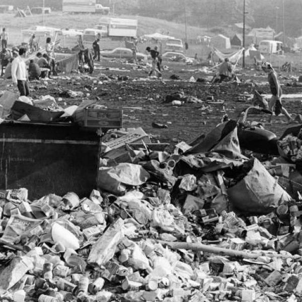 A black and white photo of the afternath of woodstock. There are piles and piles of garbage in what was once a field but is now mud