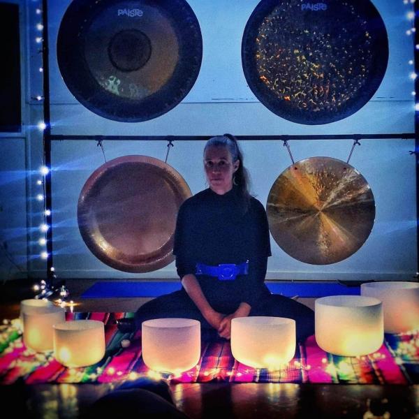 Erika Grant sits cross legged in front of four large gongs that are hanging from a square frame. In front of her there are crystal meditation/prayer bowls that are aglow with warm light
