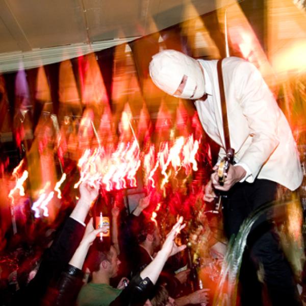 A guitarist wears a white suit, but their head is wrapped in a mask like a Mummy. There is an enthusiastic crowd raising their hands and reaching out in admiration