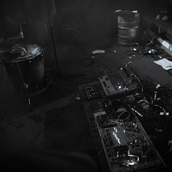 A messy and intriguing set up of cables and boxes and lights. A make shift noise synth. It is in bold black and white. There is a filthy carpet, an empty beer keg, and a dismantled drum kit, in the background.