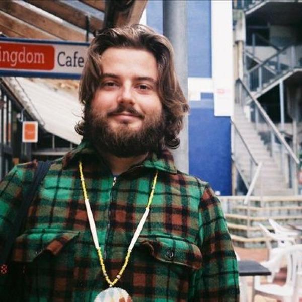 Zak is standing in the left bank off Cuba St. There are signs for restaurants, and outdor tables. Zak is wearing a warm bush shirt, and has a thick beard. He smiles gently through his whiskers.