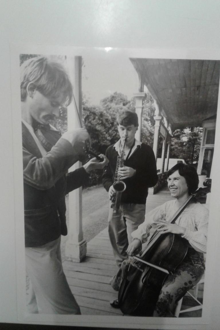 A trio plays on a verandah; Peter on viola, next is Harry Sinclair on alto sax, and then Pam Gray is playing cello. The two standing have their eyes closed in concentration, while Pam has her eyes open and is looking to Peter with a smile.