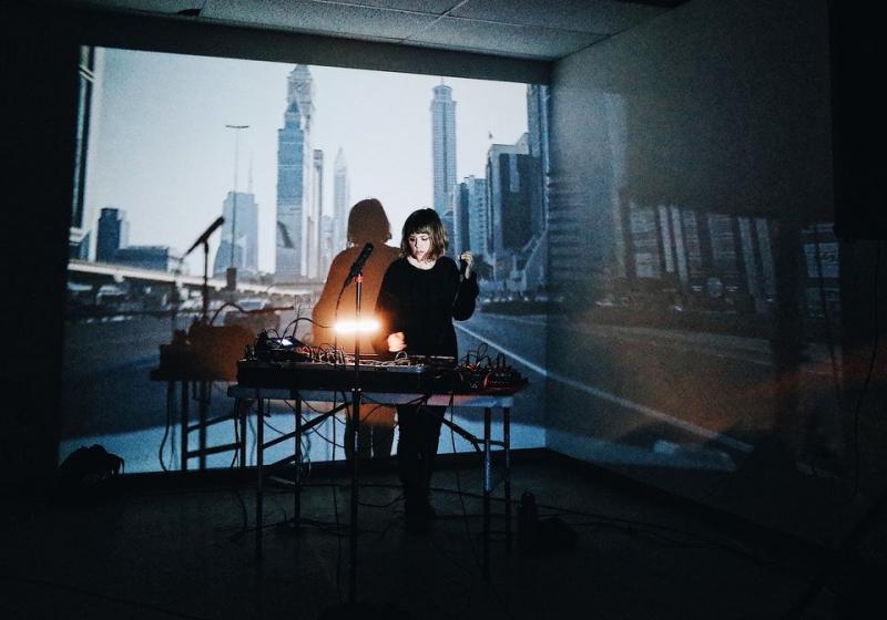 Jess Playing live, an array of electronics in front of her, and a city scene projected on the wall behind her.