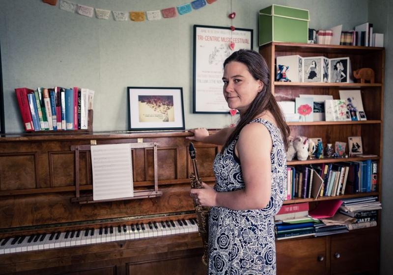 Jasmine standing in front of an an old upright piano. It is a very doemnstic setting with photos on top of the piano and a pleasingly unitdy bookshelf in the background.