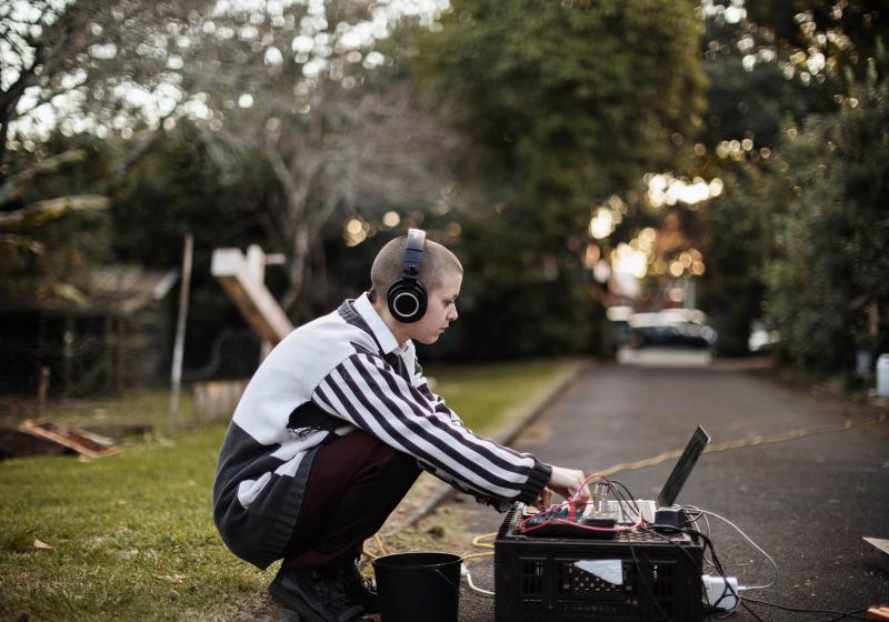 Tash leans over a laptop making a field recording in a tree lined park.