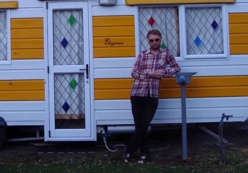 Mark is leaning nonchanontly in front of a happy yellow and white caravan
