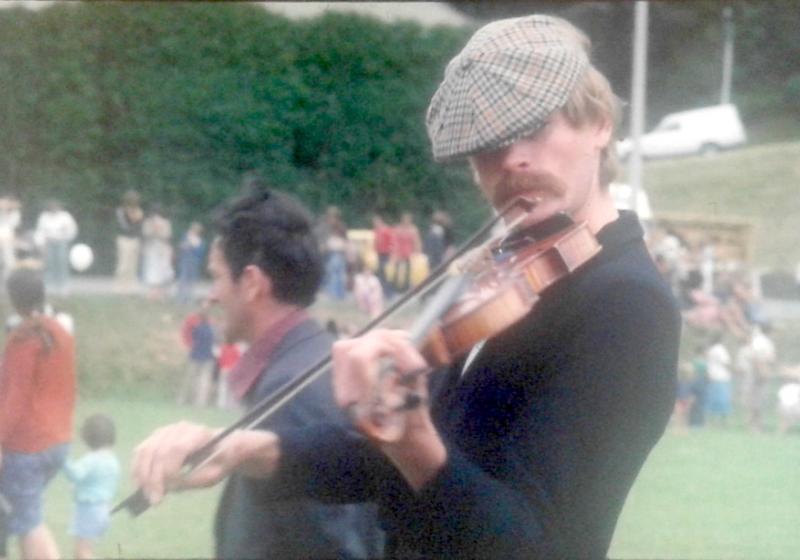 Peter playing viola outdoors. He has a robust moustache and is wearing a cheese cutter hat.