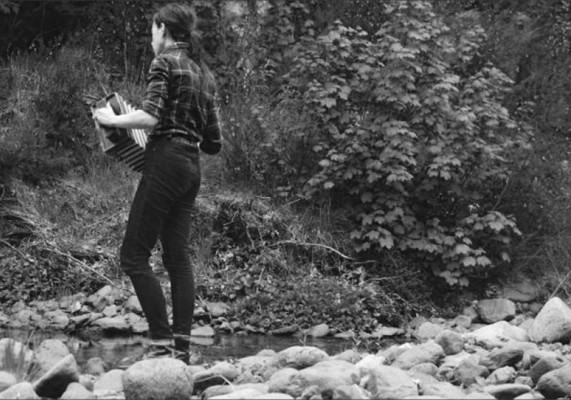 Sally is facing away from the camera. She is on the dry rocks beside a river. She is playing an accordian