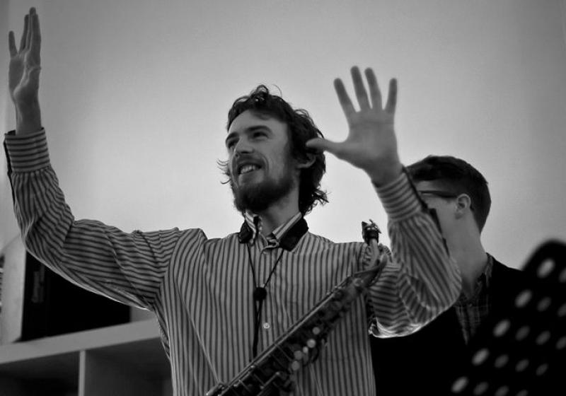 A back and white photo of Jake holding his hands up in a gesture of conducting some musicians unseen. There is a music stand to one side of him.