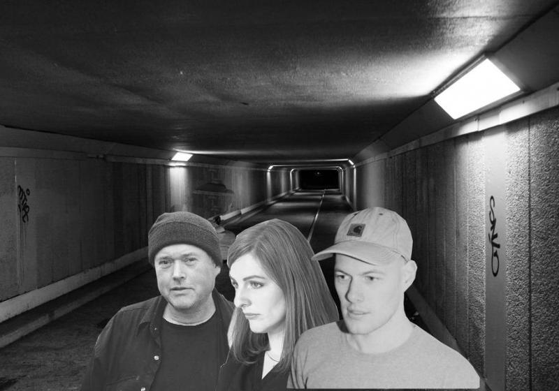 A black and white photo of the pedestrian tunnel under Wellington Airport. Collaged on top are the head and shoulders of Amy, Cory, and Omit.