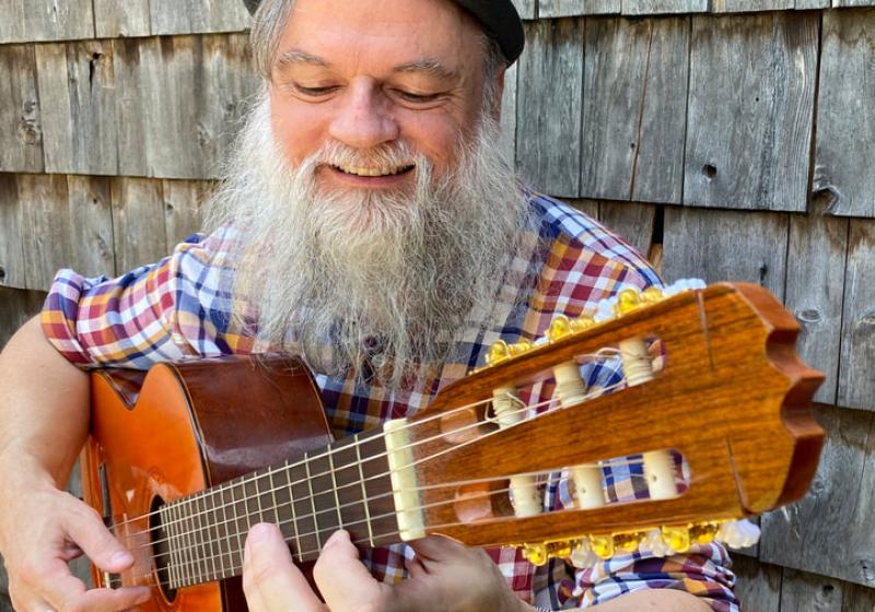 Chris has a large grey beard and is wearing a small pork pie hat. He is playing a nylon string guitar and is smiling with a large joyful smile.