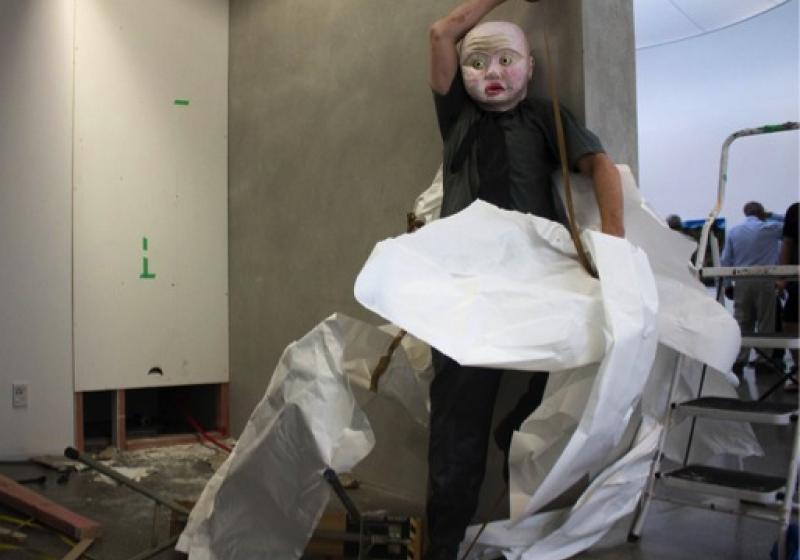 a person in a papier mache mask is leaning against a wall. They are covered in scrunched up piles of paper and they are taping themselves with brown packing tape