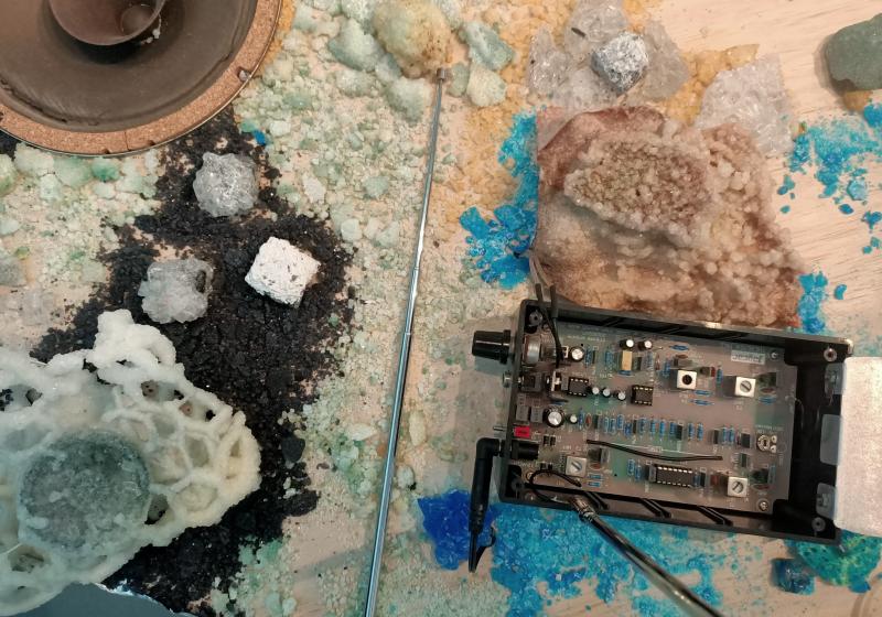 A table with whote a blue (copper?) crystals growing on it. There is a raw speaker cone and an open electronic device.