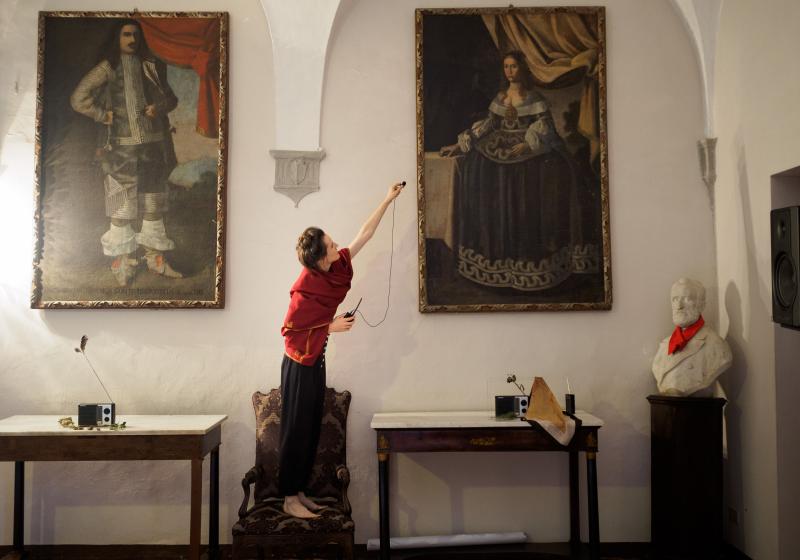 Celeste stands on a chair hanging a microphone in a gallery with classic oil paintings. Photo by Marco Giugliarelli for Civitella Ranieri Foundation