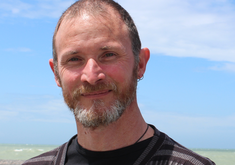 A portrait of Phil. He has a short greying beard and a gentle smile. The sky is blue, with wisps of cloud. The photo has a light feeling.