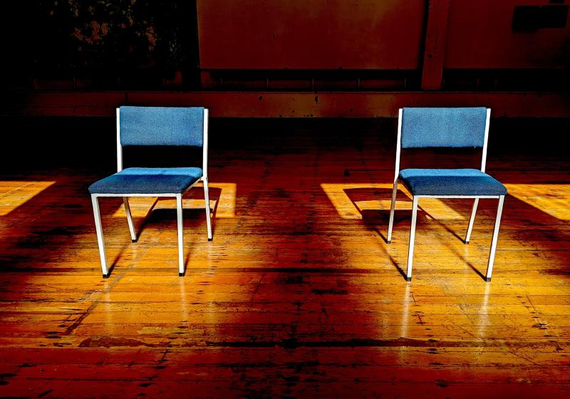 two empty chairs on in a sunbeam. The floor is wood and is possibly in a school hall.