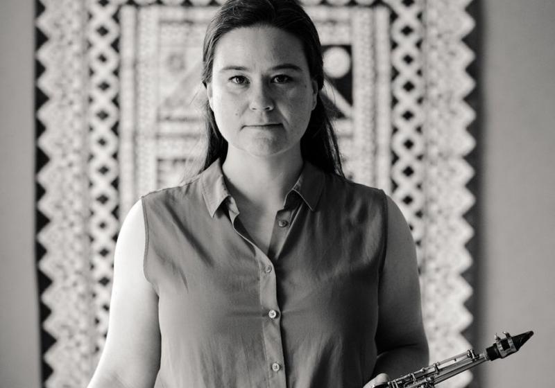 A black and white photo featuring Jasmine Lovell-Smith standing in the centre holding a soprano saxophone with a patterned tapestry hanging in the background