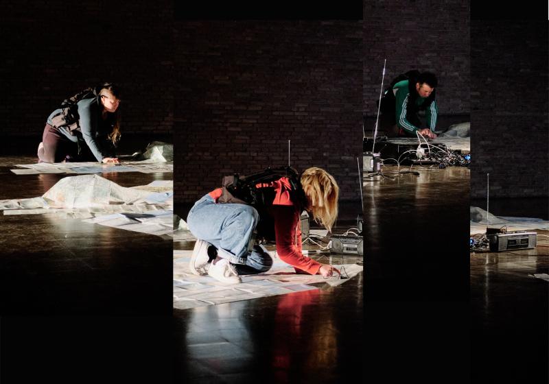 LTK4 performing on the floor of a dark stage with 1980/1990s radios, sheets of paper draped over the floor. The performers appear in work vests strapped around their torsos. 