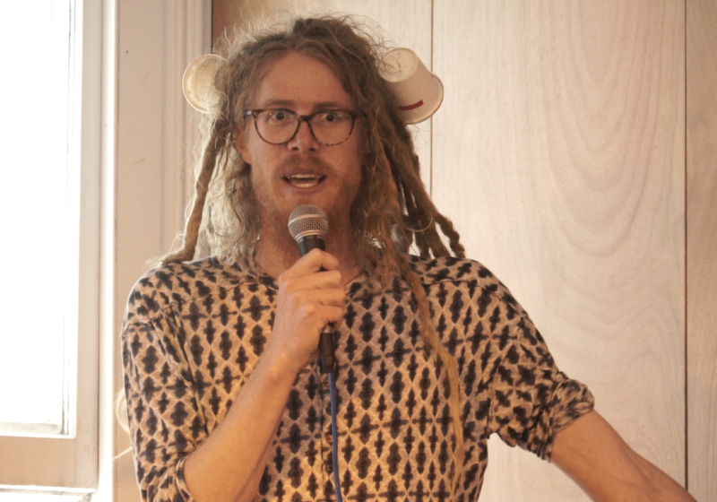 Liam has blond dreadlocks. He is wearing a patterned shirt and a pair of tortoise shell glasses. He also has two plastic cups on his head like a pair of animal ears. His eyes are open wide and his face is looking excited. He is speaking into a microphone.