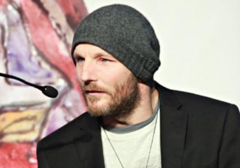 Drew is wearing a suit jacket, tshirt, and has a beanie. He is talking into a microphone that has the look of proffesionalism found at a conference or institution.