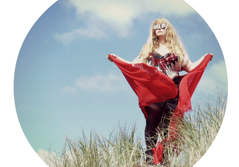 Beth is holding a red flowing skirt. She has a top covered in glitter, and sunglasses that are covered in jewels making future insect eyes. She is atop a tussock covered sand dune.