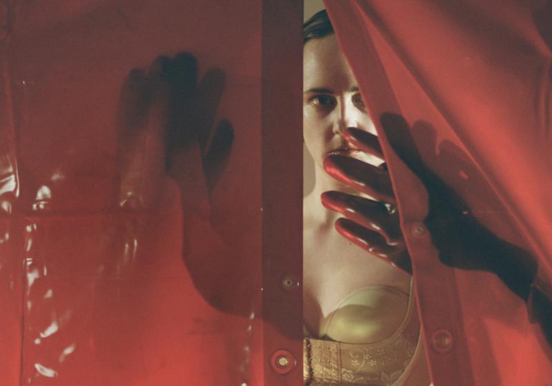 Frances in a flesh coloured corset peers through wet red transparent vinyl curtains.