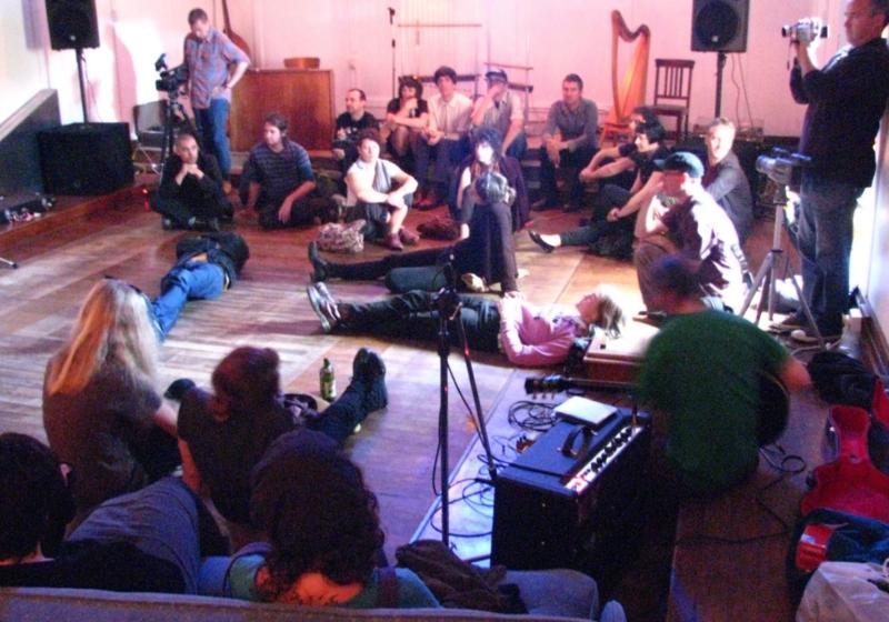 a still from the 'Lines of Flight' documentary. Audience members sit in a circle listening carefully. Some lie with their back on the floor, legs spread into the stage area in the middle of the circle.